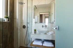 A modern bathroom with a glass-enclosed shower, a marble-tiled wall, a white sink with a vessel basin, a mirror, and a towel rack. - Theasea Stylish Residences (Mylopotamos)