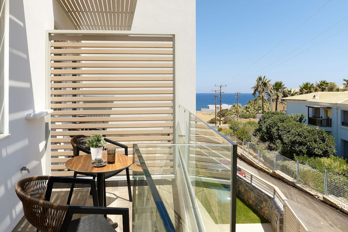 A balcony with a glass railing, a table with chairs, a potted plant, and a view of the sea and buildings.