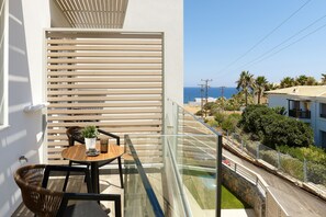 A balcony with a glass railing, a table with chairs, a potted plant, and a view of the sea and buildings. - Theasea Stylish Residences (Mylopotamos)