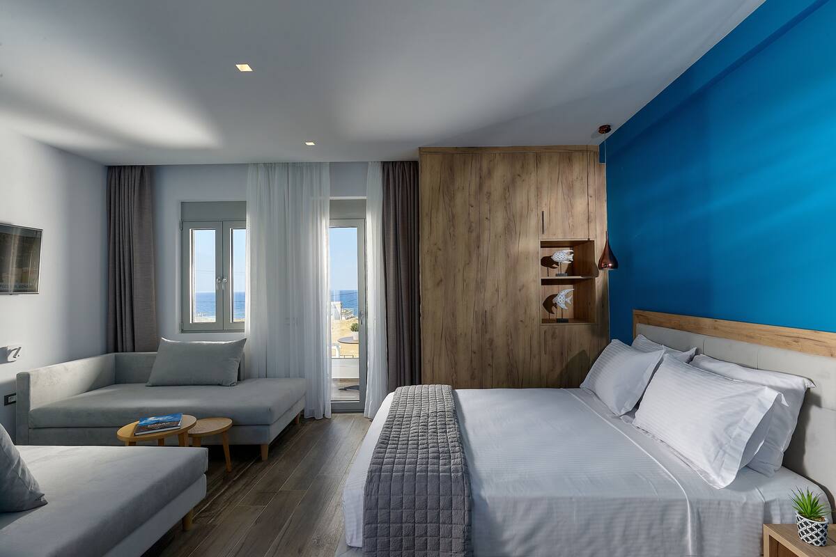 A modern hotel room with a bed, a sofa, a small table, and a view of the outdoors.