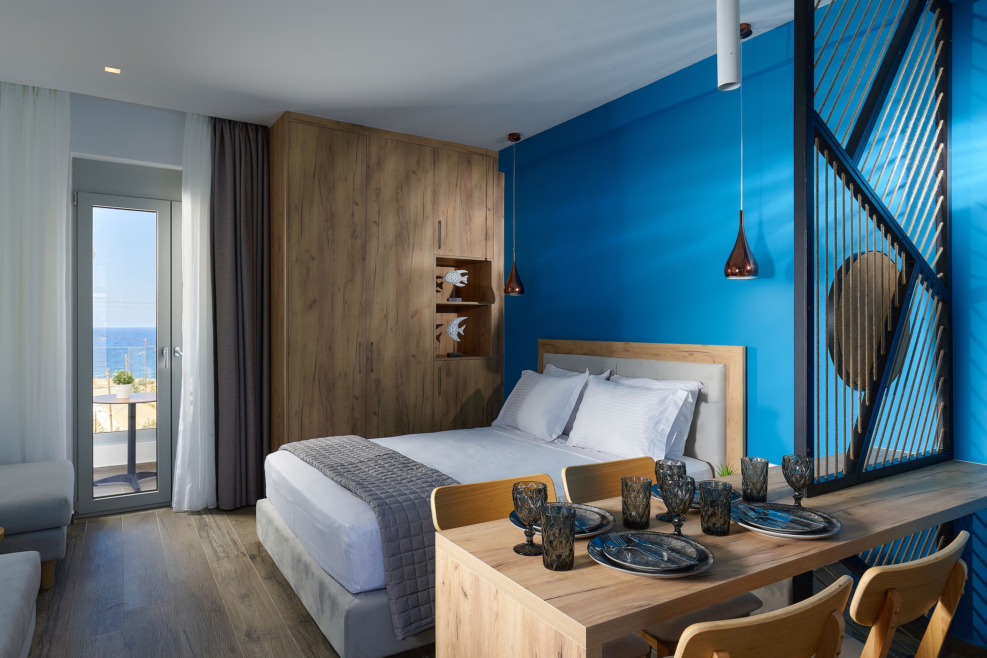A modern hotel room with a large bed, a wooden desk with dining set, and a view of the sea through a sliding door.