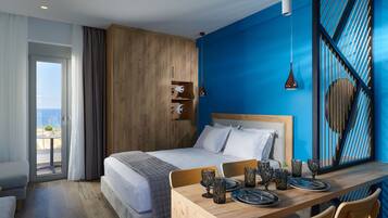 A modern hotel room with a large bed, a wooden desk with dining set, and a view of the sea through a sliding door.