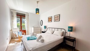 Un lit bien fait, avec du linge de lit et des serviettes blanches, une table de chevet avec une lampe, une chaise avec un coussin bleu et une fenêtre avec des rideaux.