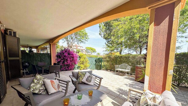 Une terrasse extérieure dotée d’un espace salon couvert, d’une table avec des boissons et offrant une vue sur la végétation et les bâtiments environnants.