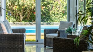 A wicker lounge chair with cushions, a glass table with a vase, and a view of a swimming pool and greenery. - Luxurious Villa "Las Hortensias" with Private Pool, Garden & Wi-Fi (Santa Brígida)