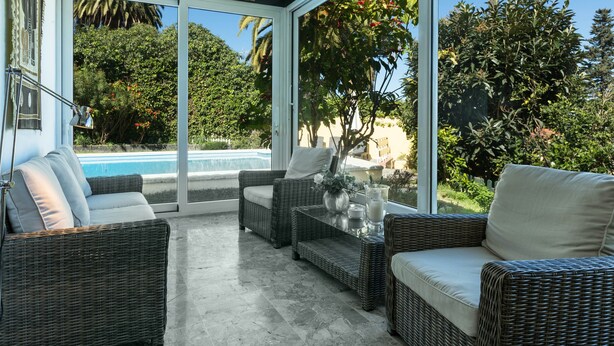 A sunlit patio with wicker furniture, a glass door leading to a pool, and lush greenery. - Luxurious Villa "Las Hortensias" with Private Pool, Garden & Wi-Fi (Santa Brígida)
