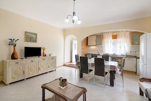 Una sala de estar con televisor, una mesa de comedor con sillas, una zona de cocina con armarios y un pasillo.
