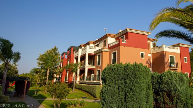 Un edificio de varios pisos con balcones, rodeado de jardines cuidados y palmeras.