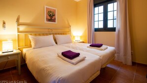 Habitación de hotel con dos camas, una mesita de noche con lámpara, una ventana con cortinas y un termostato en la pared.