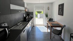 A modern kitchen with dark cabinetry, a built-in oven, and a coffee machine. There is a dining area with a wooden table and chairs, and a view of the outdoors through a door. - Ferienwohnung Weidenblick (Barth)