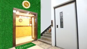 A hotel entrance with a green artificial grass wall, a wooden door frame, a welcome mat, and a staircase leading to a white door with a window. - Hotel Gaurishree (Bhopal)