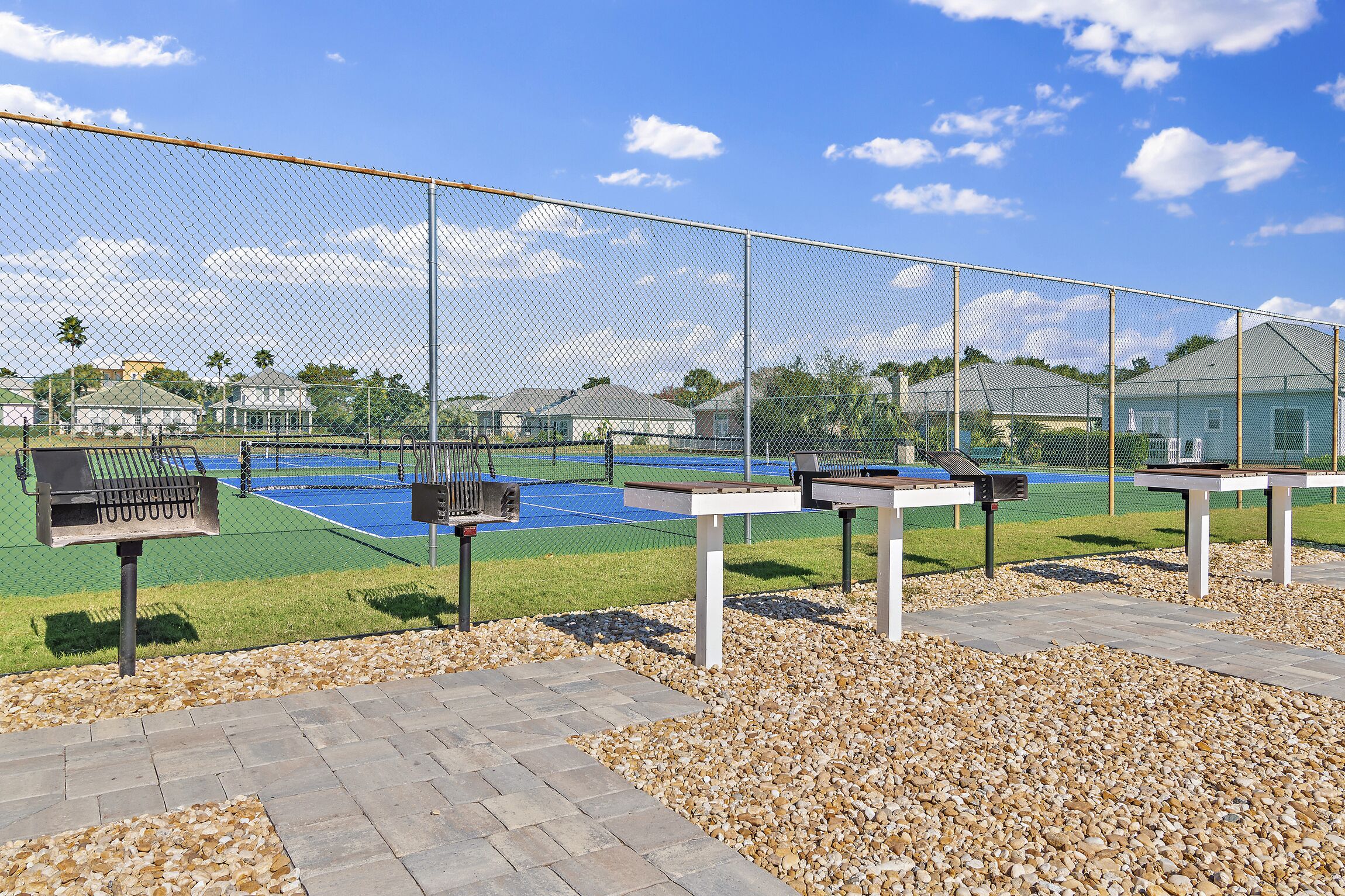 A tennis court with a chain-link fence, picnic tables, and barbecue grills.