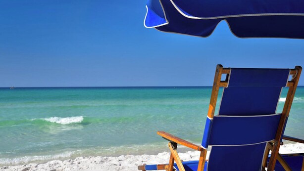Une chaise de plage et un parasol sur une plage de sable fin, avec une eau turquoise cristalline et un ciel dégagé.