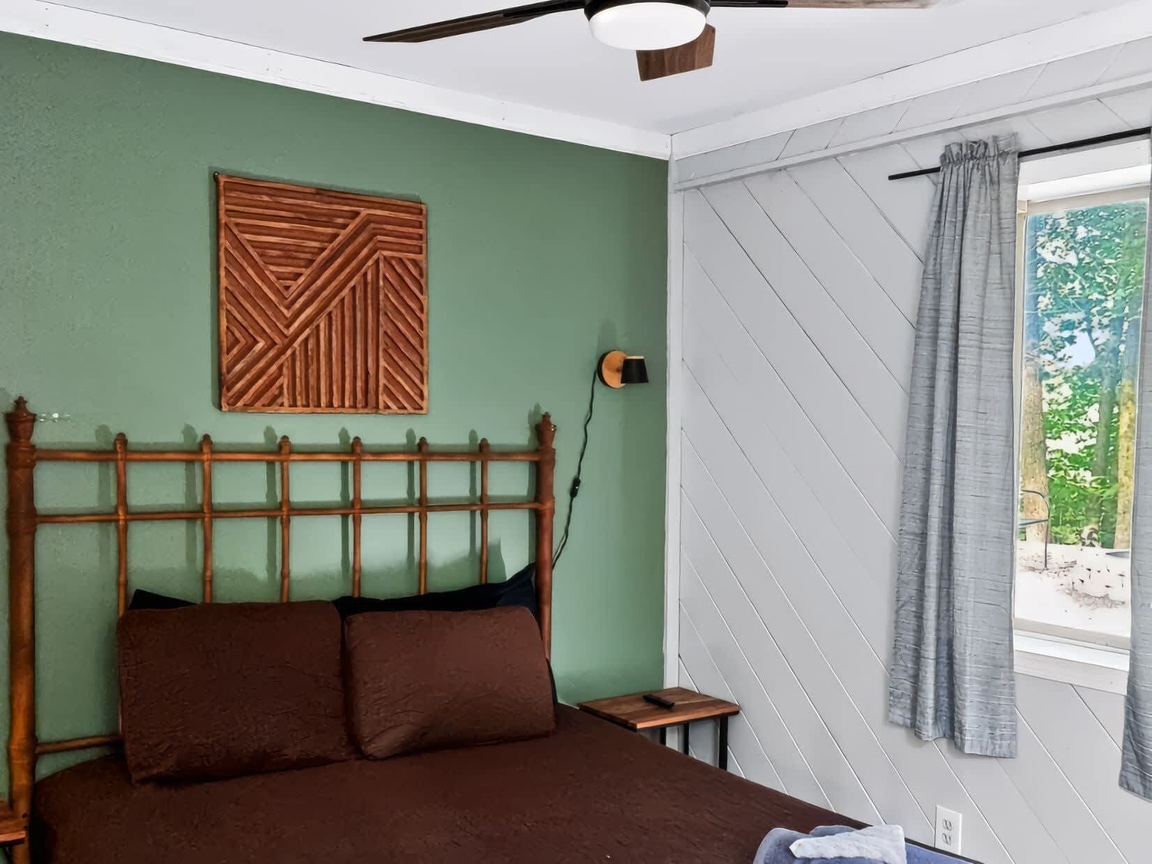 Ein Schlafzimmer mit einem hölzernen Kopfende, einem Bett mit brauner Bettwäsche, einem Nachttisch, einer Wandlampe und einem Fenster mit Vorhängen.