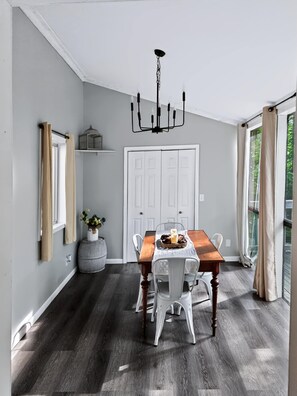 Un coin repas avec une table en bois, des chaises blanches, un lustre et une porte blanche en arrière-plan.