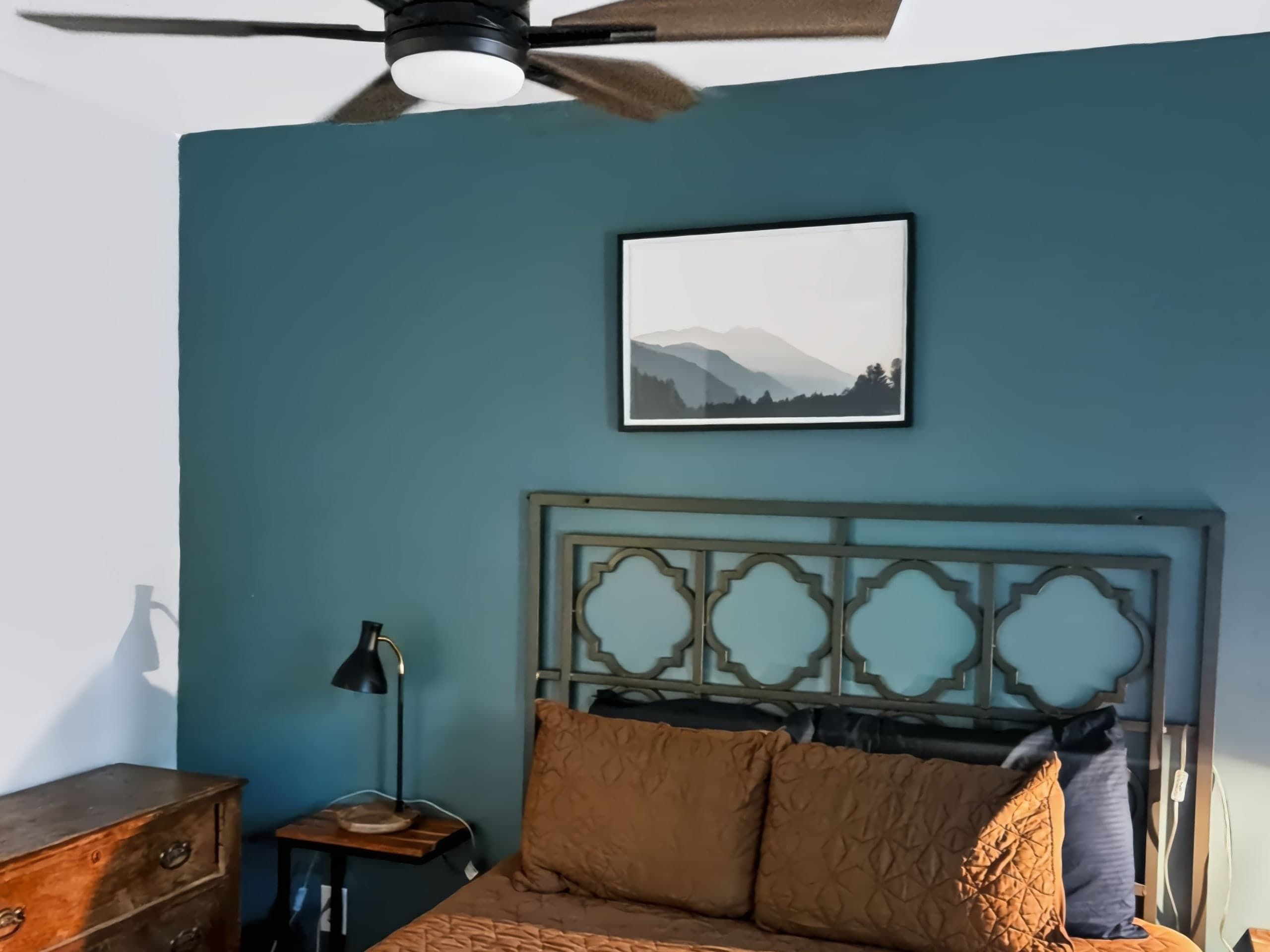 Een slaapkamer met een bed, een plafondventilator, een nachtkastje, een lamp en een ingelijst schilderij aan de muur.