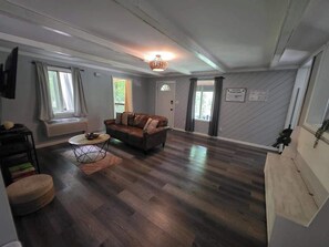 A spacious living room with a brown leather sofa, a round coffee table, and a wooden floor. There are two windows with curtains, a door, and a wall-mounted sign. - Getaway House at Avenwood Cabins (new straitsville)