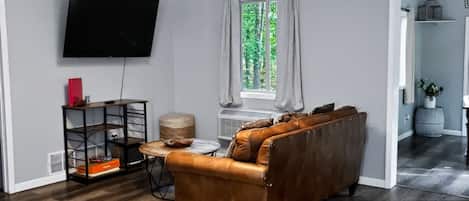 A modern living room with a leather sofa, a flat-screen TV, a wooden coffee table, and a window with curtains.
