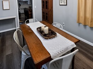 Un comedor con mesa de madera, sillas blancas, camino de mesa blanco y un centro de mesa con vela.