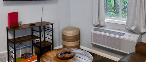 A living room with a TV mounted on the wall, a round wooden coffee table with a bowl, a leather sofa, a small bookshelf, and a window with curtains.