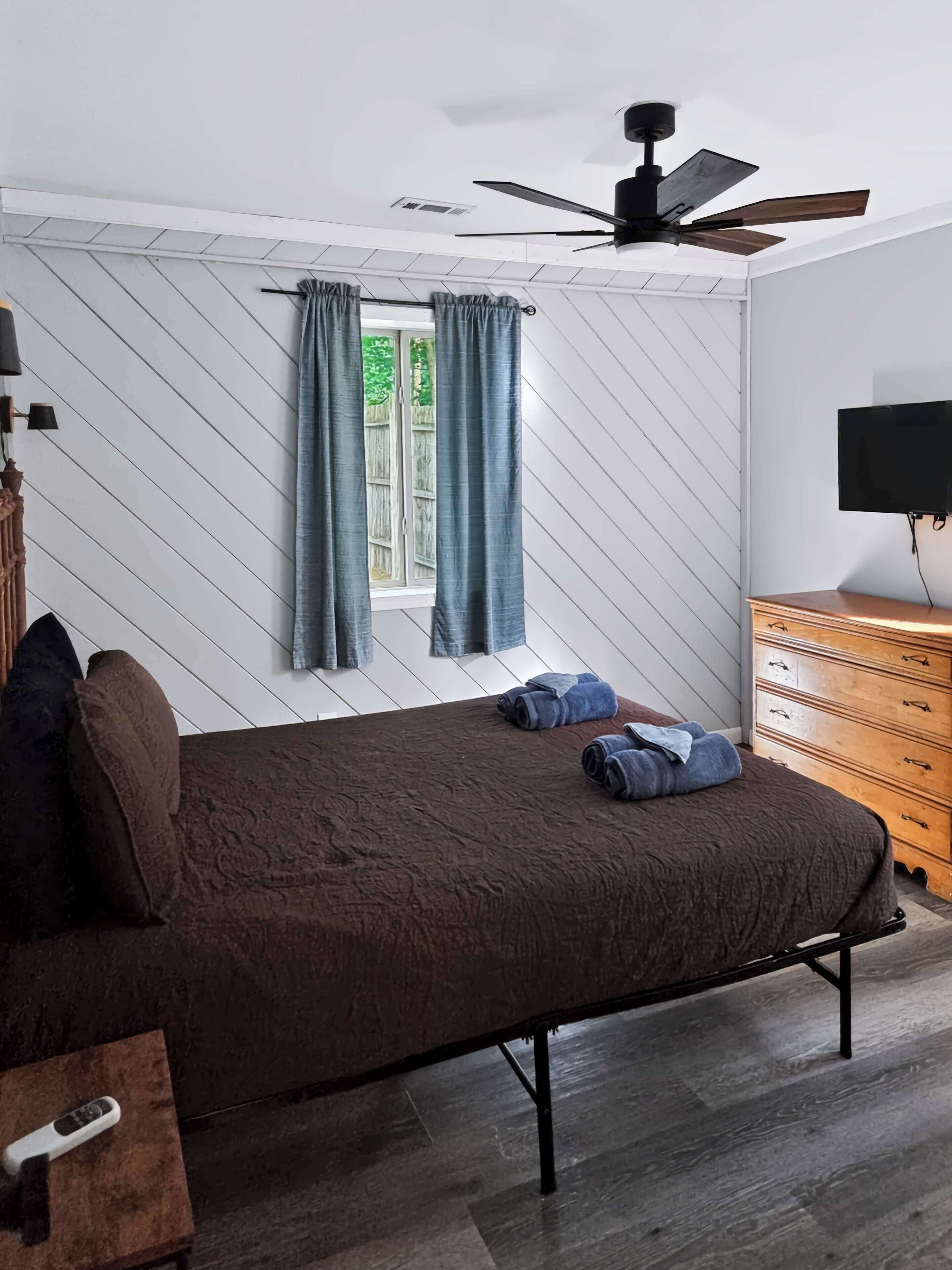 Una camera da letto con un letto, una cassettiera, una TV, una ventola a soffitto e una finestra con tende.