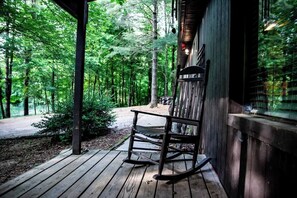 A wooden porch with a rocking chair, surrounded by a forested area.