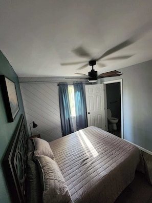 A bedroom with a bed, a ceiling fan, a door leading to a bathroom, and a window with curtains. - Getaway House at Avenwood Cabins (new straitsville)
