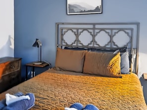 A neatly made bed with a patterned bedspread, a headboard with a decorative design, a bedside table with a lamp, and a wall-mounted picture.