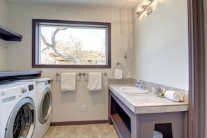 A compact bathroom with a washing machine, a sink, and a window with a view of trees and buildings. - Quiet Sea Point Cottage by Redawning (Rockport)