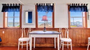 Une pièce avec trois fenêtres, chacune ornée d’une lambrequin bleue. On y trouve une table blanche et deux chaises placées devant.