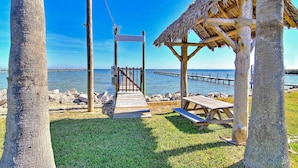 Une terrasse en bois mène à une construction au toit de chaume, située au bord d’une mer d’un bleu limpide, avec une jetée qui s’avance dans l’eau.
