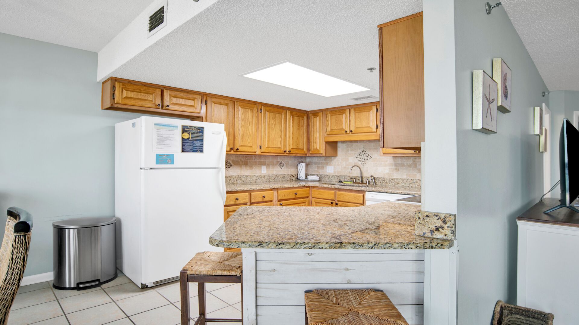 A compact kitchen with wooden cabinets, a granite countertop, a stainless steel refrigerator, and a small trash bin.