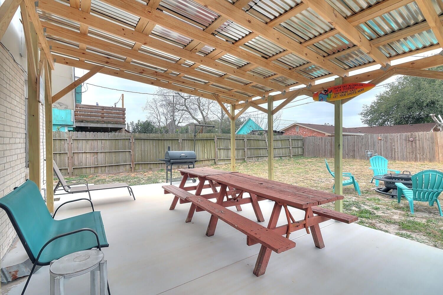 A covered patio with a picnic table and chairs, a grill, and a wooden fence in the background.