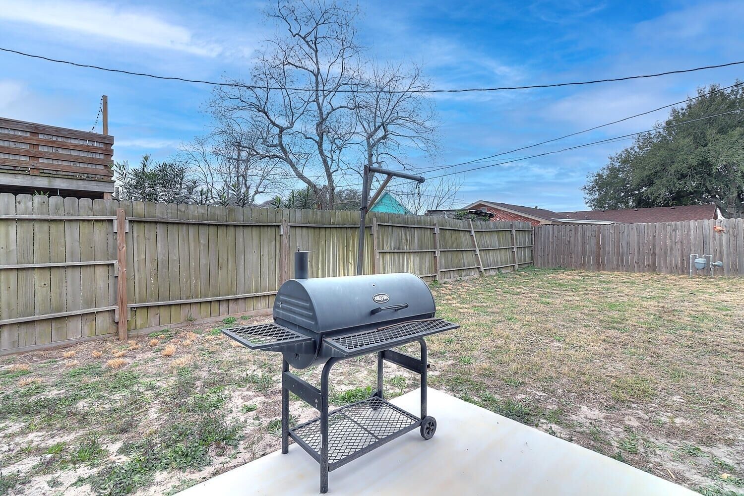 Un barbecue dans un jardin, avec une clôture en bois, un arbre et une maison en arrière-plan.