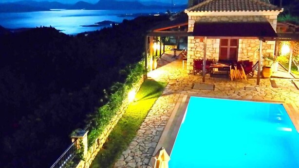 A stone building with a tiled roof overlooking a body of water, a swimming pool, and a stone pathway leading to a seating area. - Executive Villa Madouri With Private Pool (Lefkada)