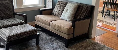 A living room with a sofa, two armchairs, a patterned rug, a wall-mounted air conditioner, and a sign on the wall.