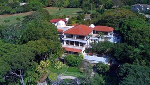An aerial view of a large estate with multiple buildings, including a main house with a red roof and a white dome structure, surrounded by lush greenery and a well-maintained garden. - Villa Elsewhere  | Ocean View - Located in  Stunning Sandy Lane with Private Pool (Holetown)
