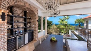 A spacious patio with a brick fireplace, a chandelier, and a view of the ocean. - Villa Elsewhere  | Ocean View - Located in  Stunning Sandy Lane with Private Pool (Holetown)