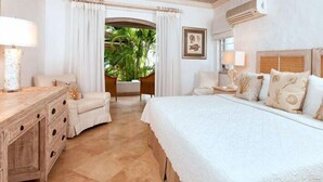 A hotel room with a large bed, a wooden nightstand, a desk, and a view of a tropical garden. - Emerald Beach 3 - Villa Ixoria Summer Promotion | Beach View - Located in  Beautiful Gibbs Bay with Private Chef Services (Lower Carlton)
