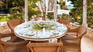 A round glass table set for six with wicker chairs, surrounded by lush greenery and a pool in the background. - Emerald Beach 3 - Villa Ixoria Summer Promotion | Beach View - Located in  Beautiful Gibbs Bay with Private Chef Services (Lower Carlton)
