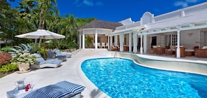 A swimming pool with lounge chairs and umbrellas, surrounded by lush greenery and a white building with a covered patio. - Villa Klairan  | Ocean View - Located in  Tropical Sandy Lane with Private Pool (Holetown)