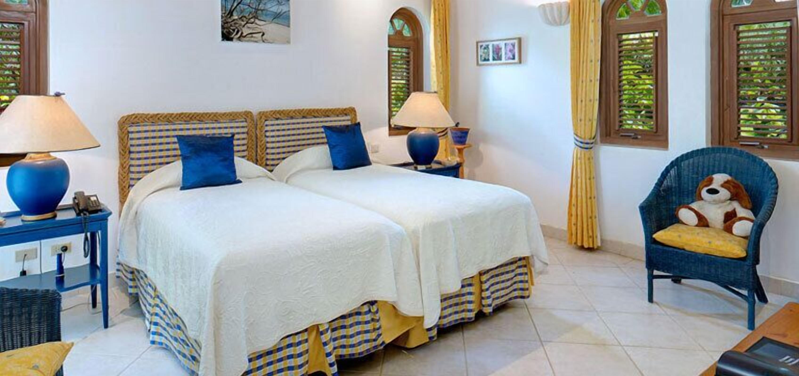 A double bed with checkered bed runners, two bedside lamps, a wicker chair with a cushion, and a small table with a lamp.
