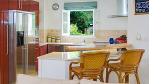 A compact kitchen with a refrigerator, a sink, and a window with a view of greenery. There are wicker chairs and a small dining table. - Schooner Bay 108 - Condo Chilterns | Beach Front - Located in  Magnificent St. Peter with Concierge Services (St. Peter)