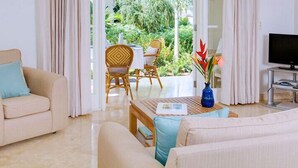 A hotel room with a sofa, a wooden table, a TV, and a view of the garden. - Schooner Bay 108 - Condo Chilterns | Beach Front - Located in  Magnificent St. Peter with Concierge Services (St. Peter)