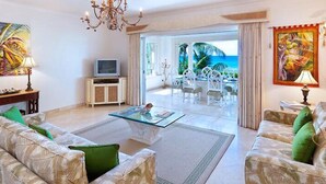 Un salon spacieux avec une table basse en verre, deux canapés avec des coussins verts, un téléviseur posé sur un meuble en bois et une vue sur l’océan grâce à de grandes fenêtres.