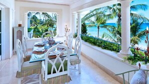 Un espace repas offrant une vue sur l’océan, comprenant une table avec des chaises, une plante décorative et une vue sur les palmiers et la mer.