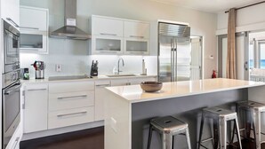 A modern kitchen with white cabinetry, a stainless steel range hood, a large island with stools, and a view of the ocean through the window.