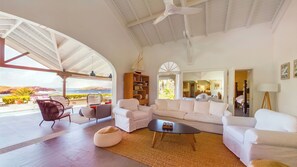 A spacious living area with a white sofa set, a coffee table, and a bookshelf. A large window offers a view of the ocean and a mountainous landscape.