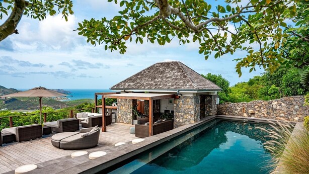 Un espace aménagé au bord de la piscine, avec un mur en pierre, un coin salon abrité et une vue sur l’océan.