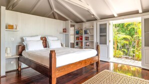 Une chambre avec un lit en bois, une table de chevet, une bibliothèque et une vue sur un jardin tropical.